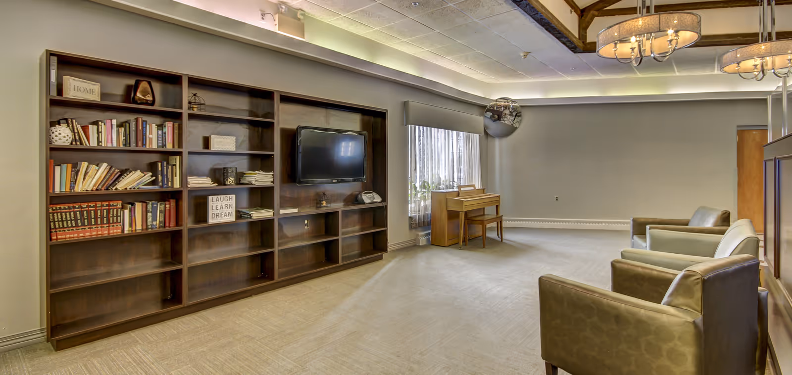 A spacious living room area with a large wooden bookshelf filled with books and decorative items, a wall-mounted flat-screen TV, a small piano near a window with sheer curtains, and three cushioned armchairs arranged facing the TV. The ceiling has recessed lighting and two hanging chandeliers.