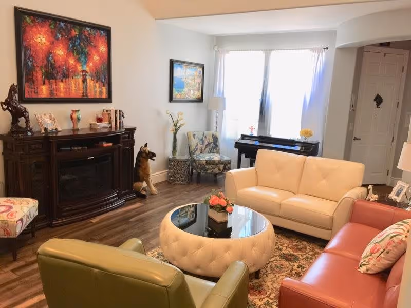 A cozy living room with a cream leather sofa, a red leather sofa, a green armchair, and a floral patterned chair. A round tufted ottoman with a glass top and a flower arrangement sits in the center on a patterned rug. There is a dark wooden cabinet with decorative items and a statue of a horse, two colorful paintings on the walls, a piano near a window with white curtains, and a door in the background.