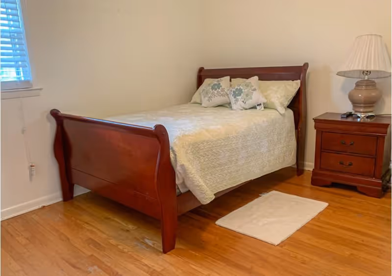A bedroom with a wooden bed frame featuring a light-colored quilt and three decorative pillows. Next to the bed is a wooden nightstand with two drawers, a beige lamp, and a remote control. The room has hardwood flooring, a small white rug beside the bed, and a window with blinds partially open.
