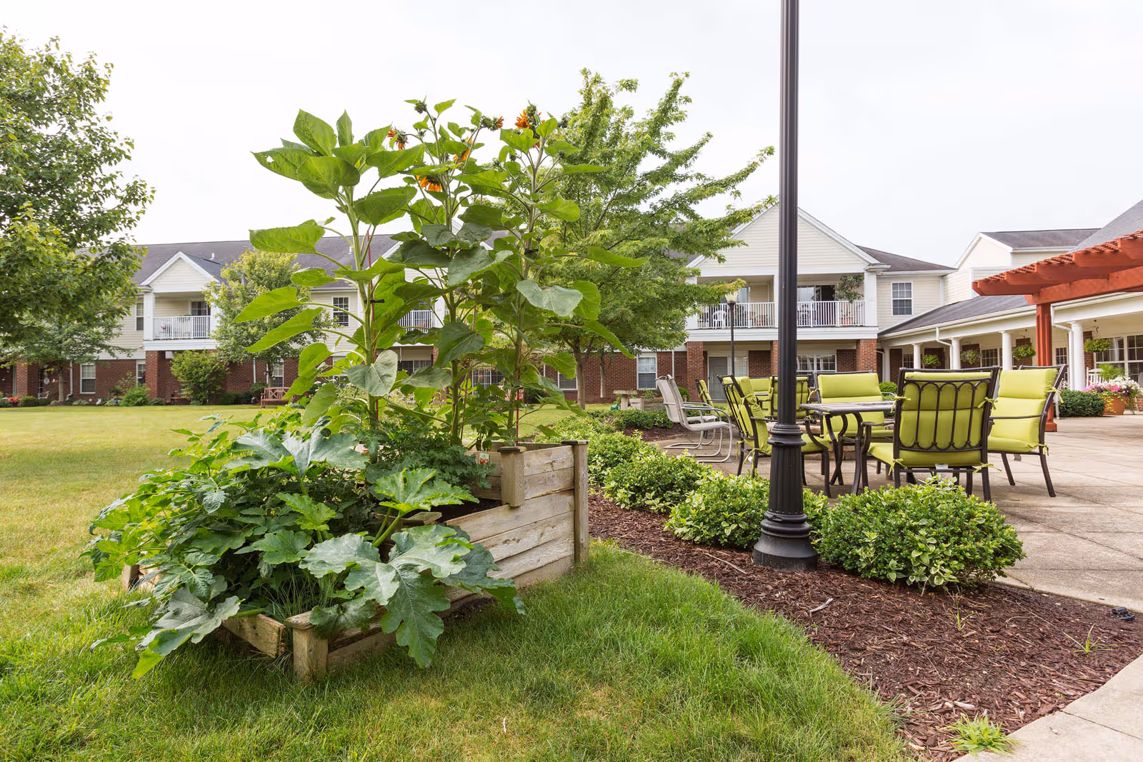 Courtyard with a raised wooden planter, patio tables and green cushioned chairs in front of a two-story senior living building.