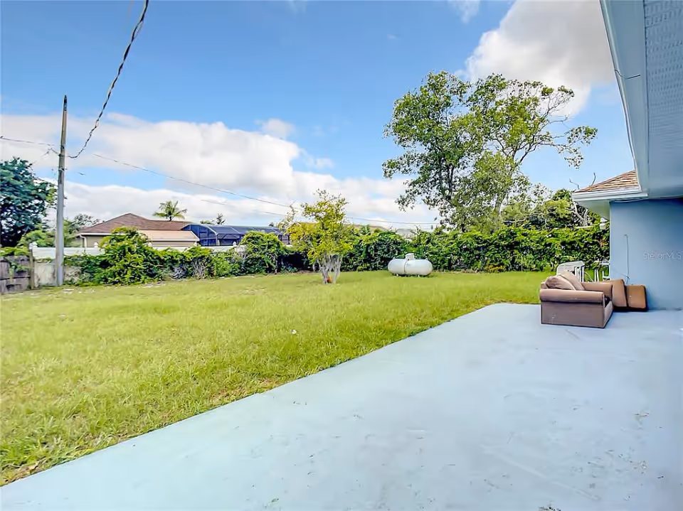 A spacious backyard with green grass, a few trees, and a clear blue sky with some clouds. There is a concrete patio area with a brown outdoor sofa set against the side of a light blue house. In the background, there are some bushes and a propane tank visible.