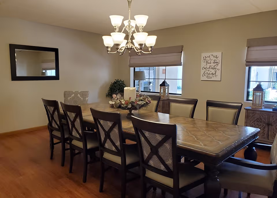 A dining room with a long wooden table surrounded by eight chairs, a chandelier with multiple lights hanging above the table, two windows with beige roman shades, a wall mirror, a decorative wreath centerpiece on the table, and a wall sign that reads 'give us this day our daily bread'.