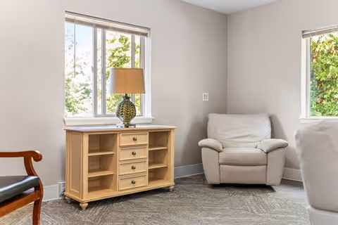 A cozy corner of a living room with two large windows letting in natural light. There is a beige armchair in the corner, a wooden cabinet with shelves and drawers, and a table lamp on top of the cabinet. Part of another chair with wooden armrests and black seat is visible on the left side.