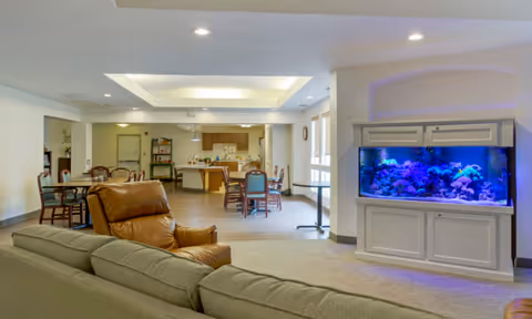 Open senior living common area with sofas and recliner, dining tables, a kitchenette in the back, and a large illuminated aquarium.