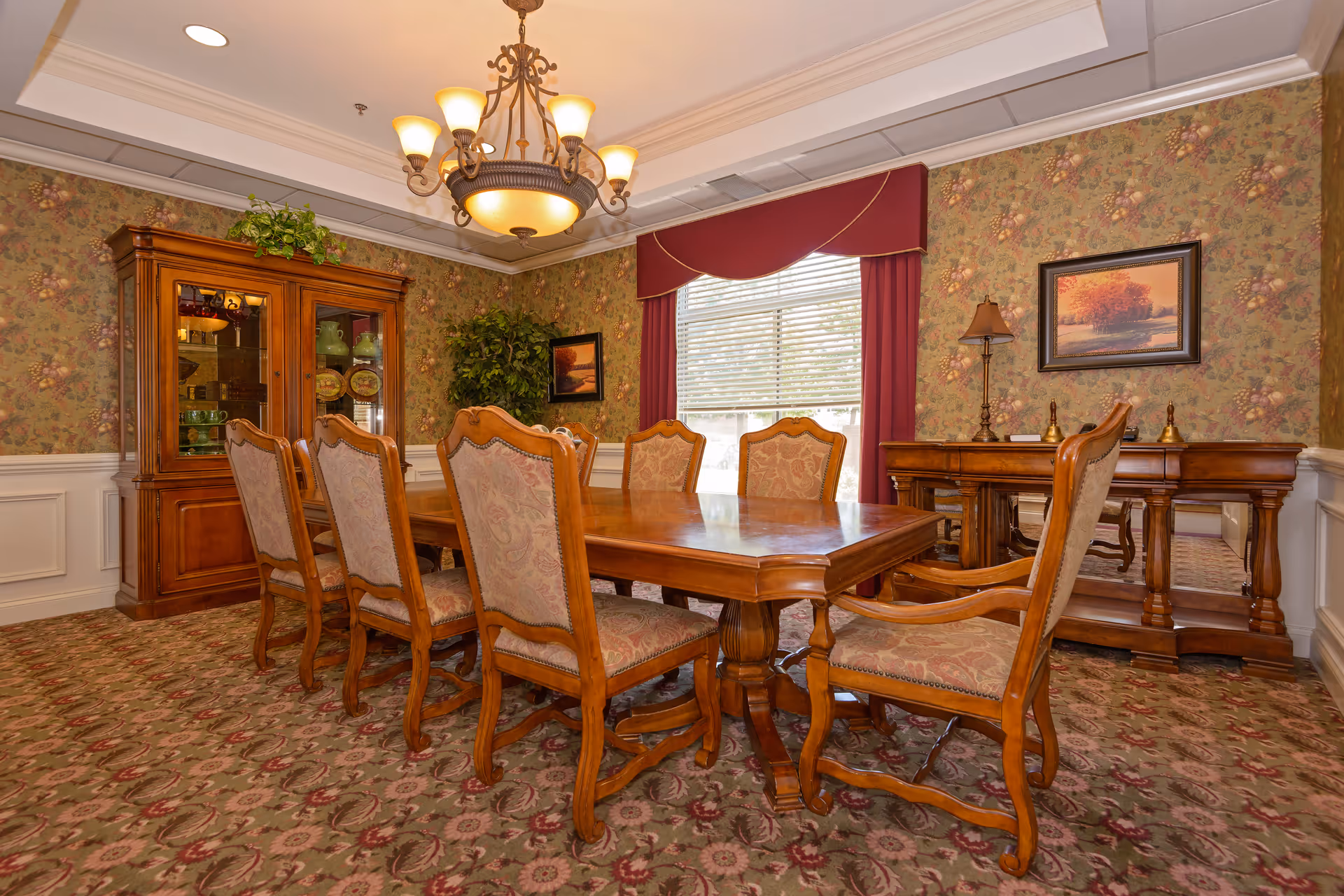 A traditional dining room with a wooden dining table surrounded by eight upholstered chairs. The room features floral wallpaper, a large window with red curtains, a chandelier hanging from the ceiling, a wooden china cabinet with decorative items, a sideboard with a lamp, and framed artwork on the walls.