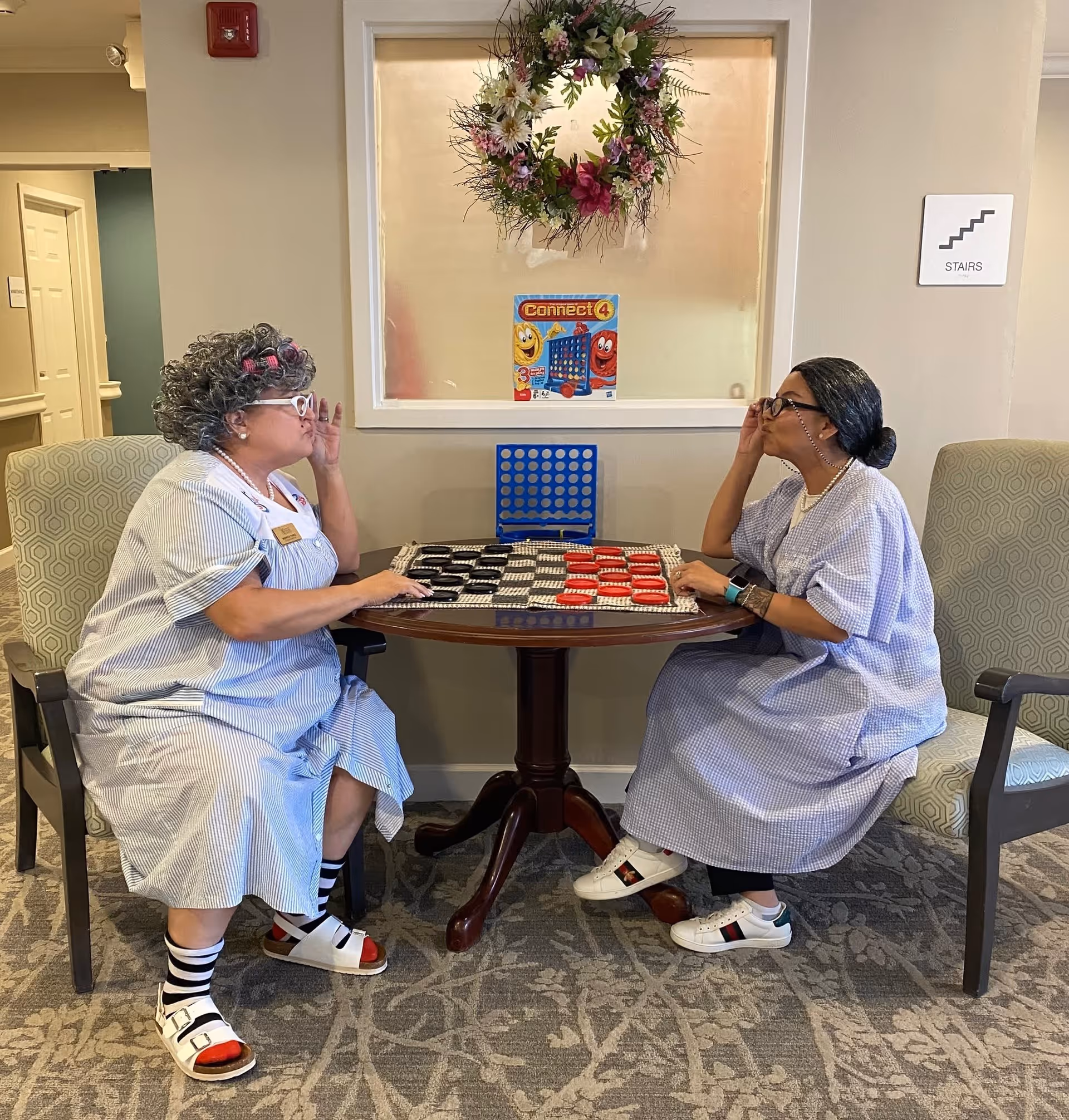 Two elderly women dressed in light blue striped dresses and glasses are sitting across from each other at a small round table playing a board game with large red and black pieces. Behind them is a window with a floral wreath hanging above a Connect 4 game box. The setting appears to be a common area in a senior living facility.