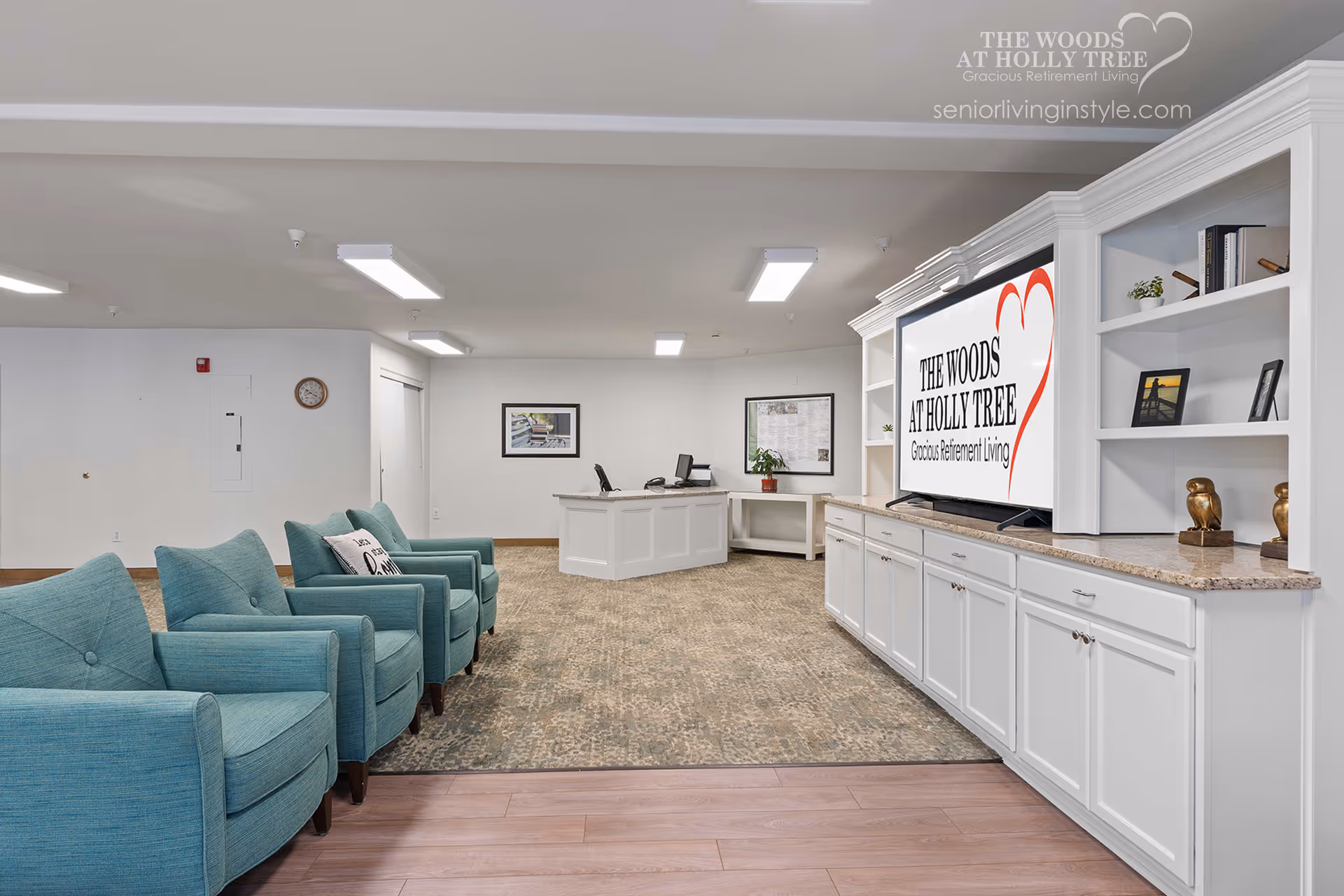 A bright communal lounge with teal armchairs facing a built-in media cabinet and TV in a retirement community.