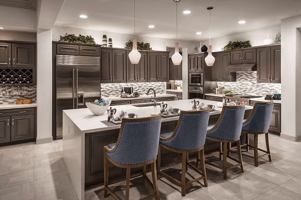 Modern open kitchen with a large white island, four blue upholstered bar stools, gray cabinetry, stainless steel appliances, and pendant lights.