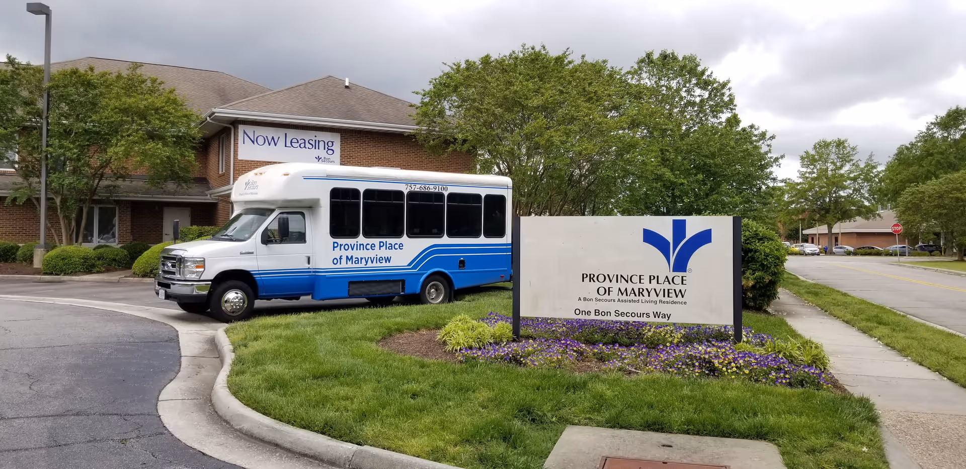Exterior view of Province Place of Maryview assisted living residence showing a white and blue shuttle bus parked near the building. A large sign with the facility's name and logo is visible in the foreground, surrounded by grass and flowers. The building is brick with a 'Now Leasing' banner displayed on the side. Trees and a street with parked cars are in the background under a cloudy sky.