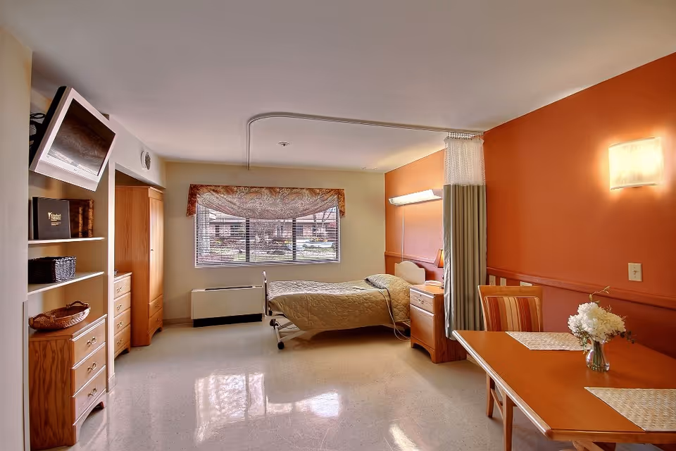 A well-lit senior care facility room with a single bed, wooden furniture including a wardrobe, drawers, and a table with chairs. The room has a large window with blinds and a patterned valance, an orange accent wall with a wall light, and a privacy curtain near the bed.