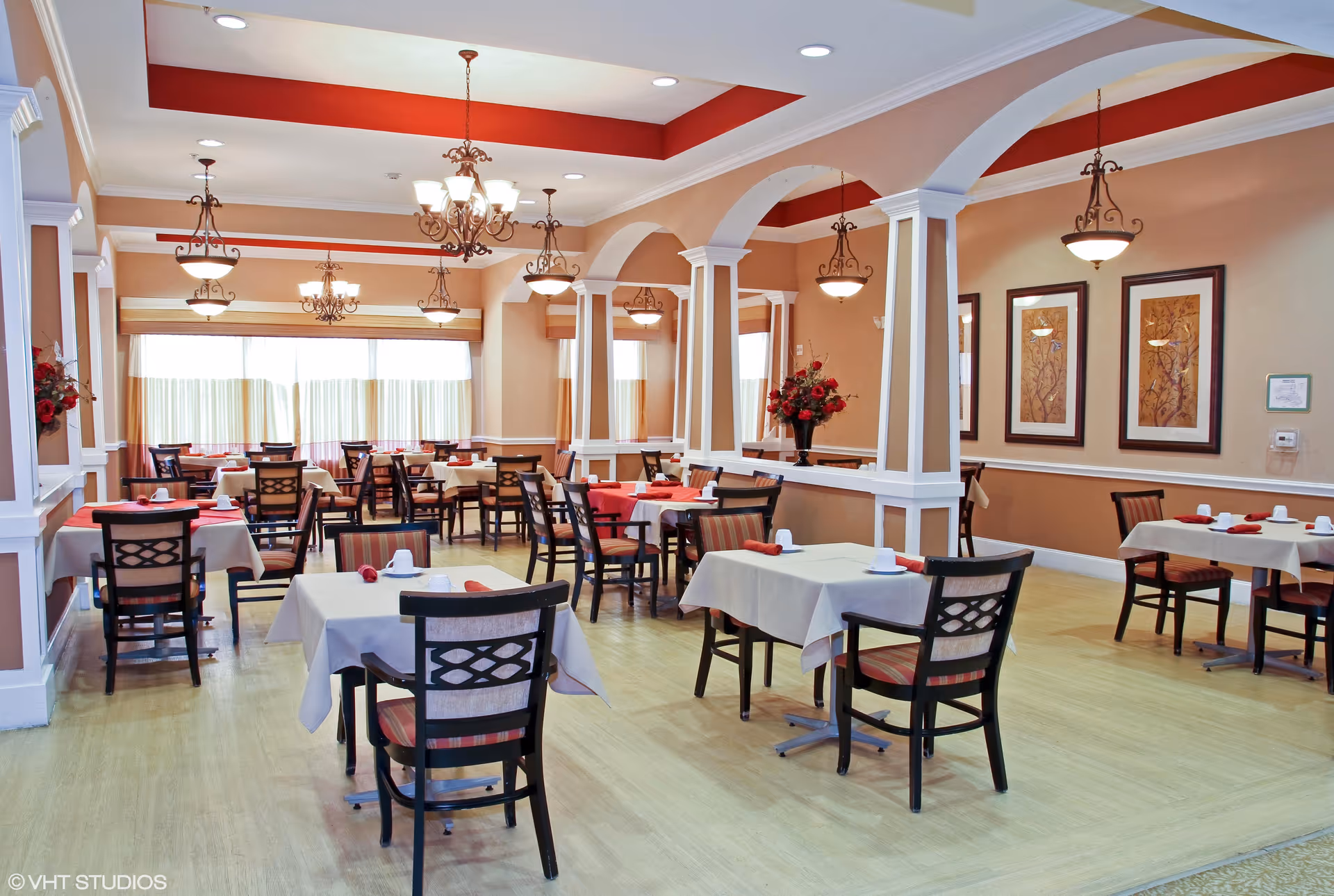 A spacious dining room with multiple tables covered in white tablecloths and set with white cups and red napkins. The room features warm beige walls with white trim, decorative columns, framed artwork, and several ornate chandeliers hanging from the ceiling. Large windows with curtains allow natural light to fill the space.
