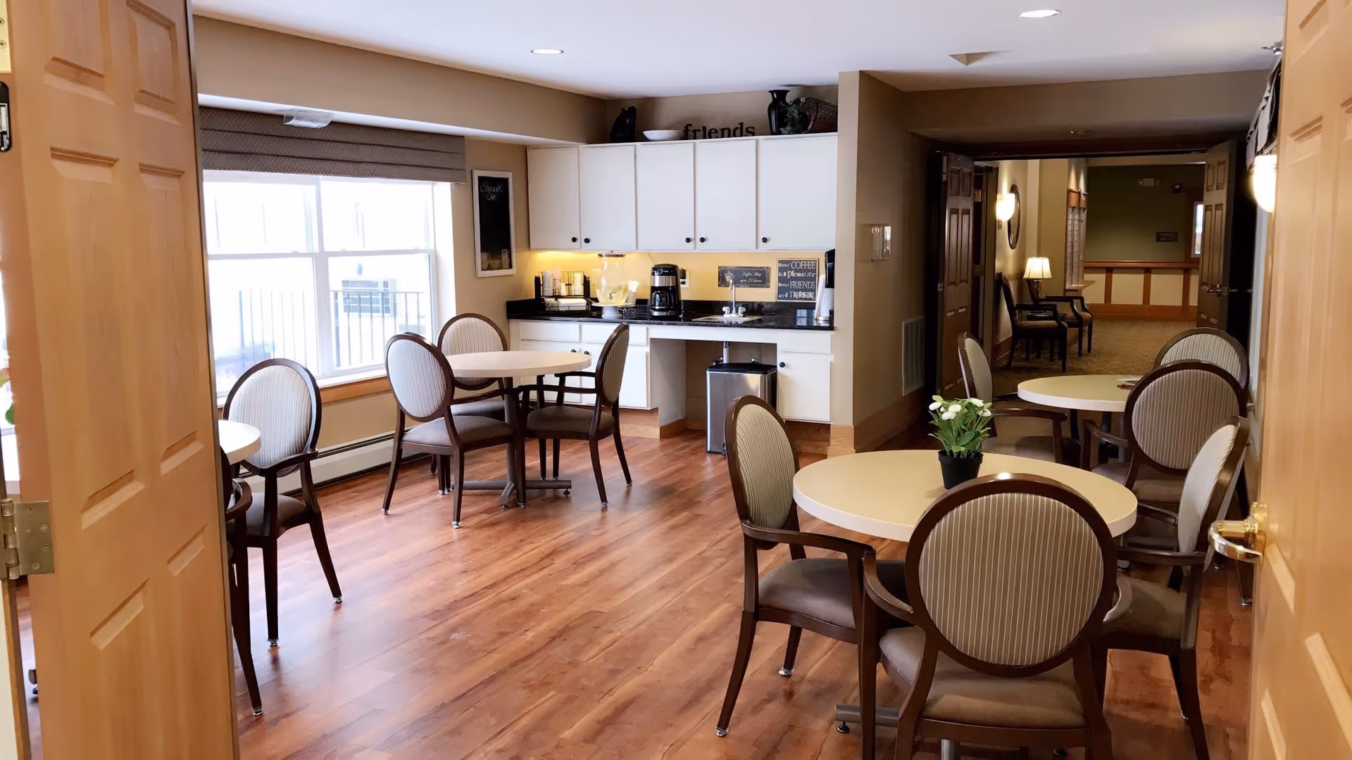 Interior view of a dining area in a senior living facility with round tables and cushioned chairs arranged on a wooden floor. There is a countertop with cabinets, a coffee maker, and a water dispenser along one wall. The room is well-lit with natural light coming through a large window and warm ceiling lights. A hallway with additional seating and lamps is visible in the background.