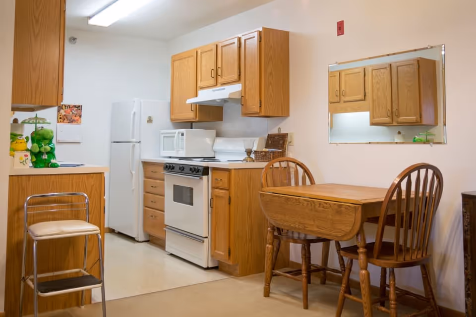 A small kitchen area with wooden cabinets, a white refrigerator, stove, and microwave. There is a wooden dining table with two chairs and a large mirror on the wall above the table. A step stool with a cushioned seat is positioned near the counter.