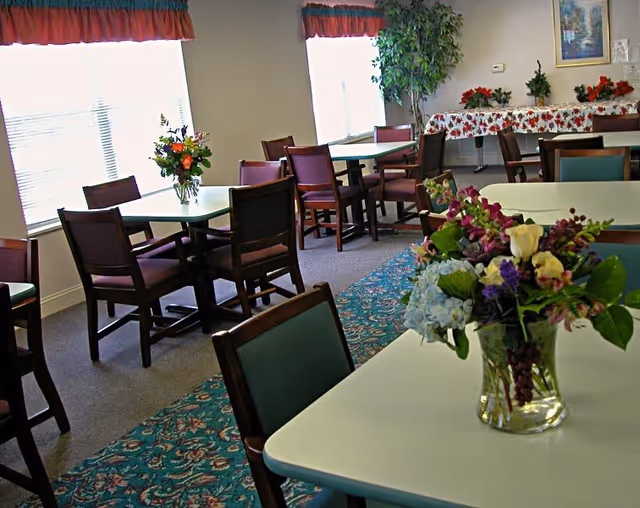 A bright dining room with several tables and wooden chairs arranged neatly. Each table has a floral centerpiece in a glass vase. The room has large windows with red valances, a patterned carpet, and a table covered with a festive tablecloth in the background. There are also some potted plants and framed artwork on the walls.