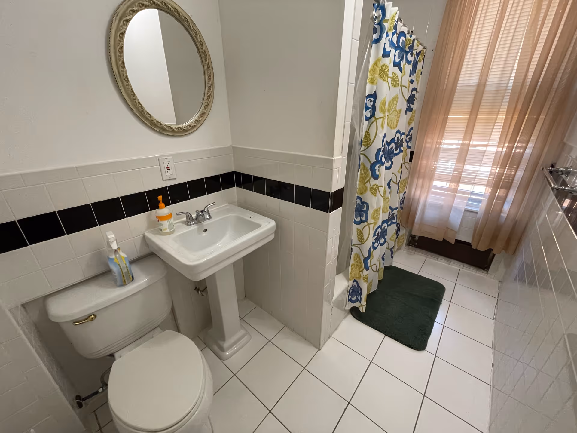 Compact bathroom featuring a toilet, pedestal sink, floral shower curtain, and sheer beige curtains.