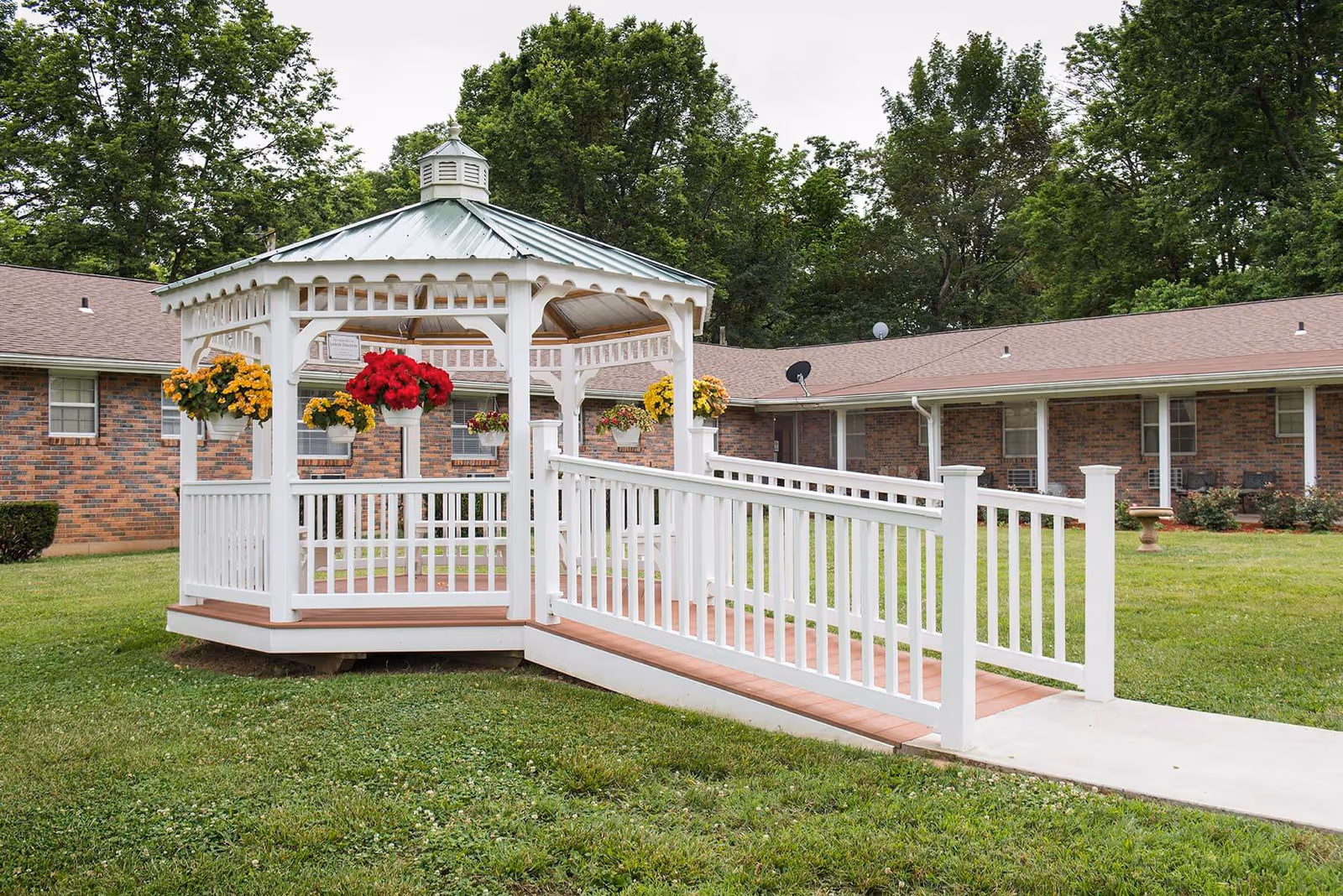 White gazebo with hanging flower baskets and a ramp in a grassy courtyard in front of a brick senior living building.