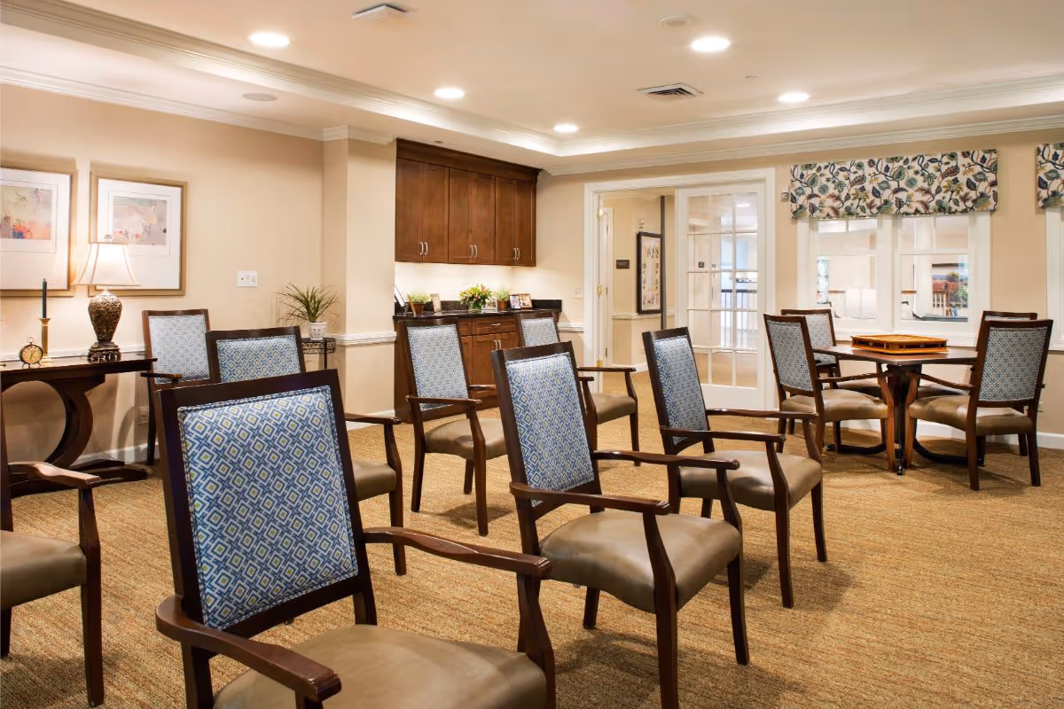 Bright communal sitting and dining area with multiple upholstered chairs arranged in rows and small tables near windows.