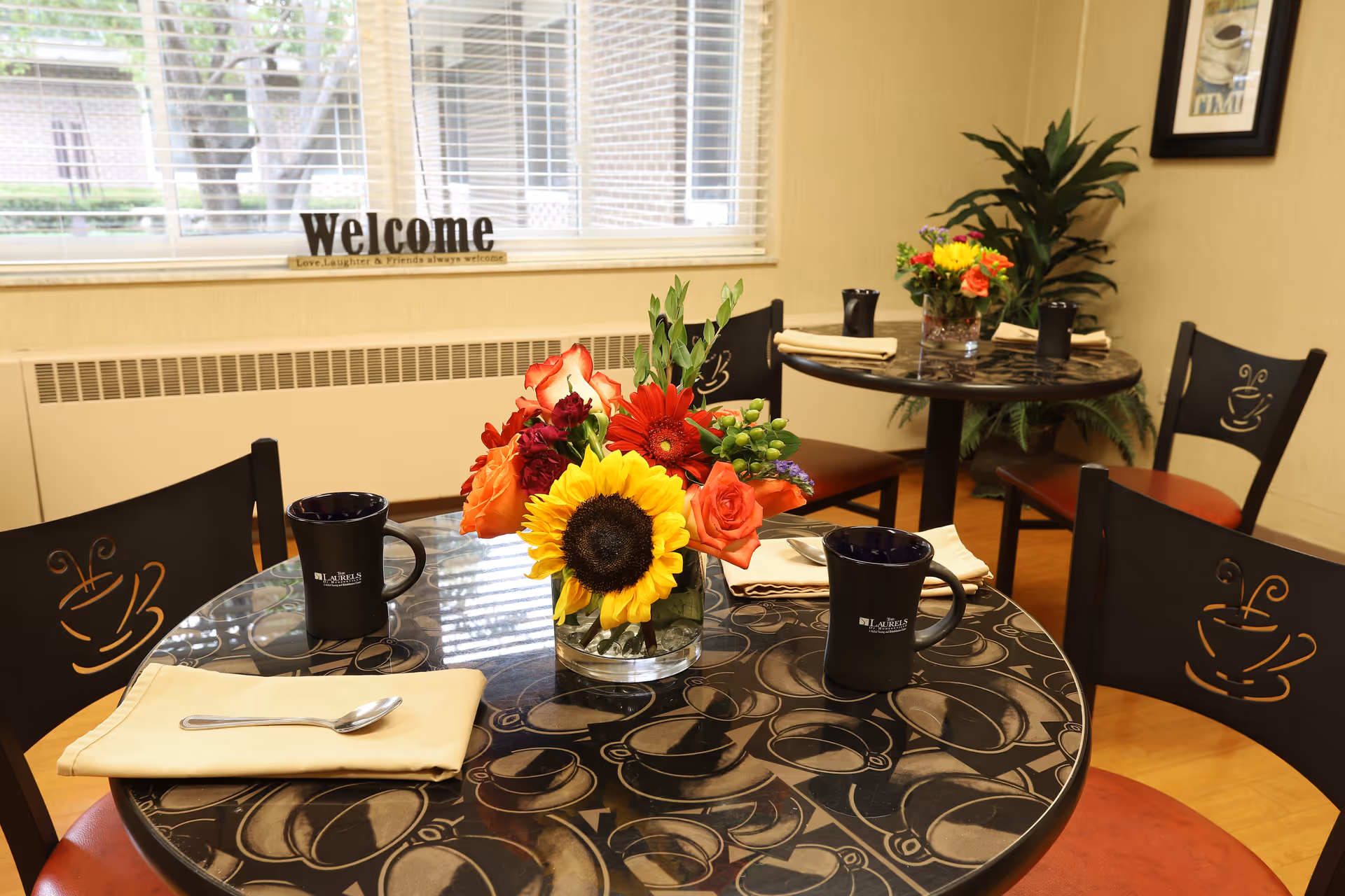 A cozy dining area with round tables featuring black and brown patterned tabletops. Each table is set with black mugs, beige napkins with spoons, and a colorful flower arrangement including a sunflower, roses, and other flowers. Black chairs with a coffee cup design on the backrest surround the tables. A window with blinds and a 'Welcome' sign is visible in the background, along with a green potted plant and framed artwork on the wall.