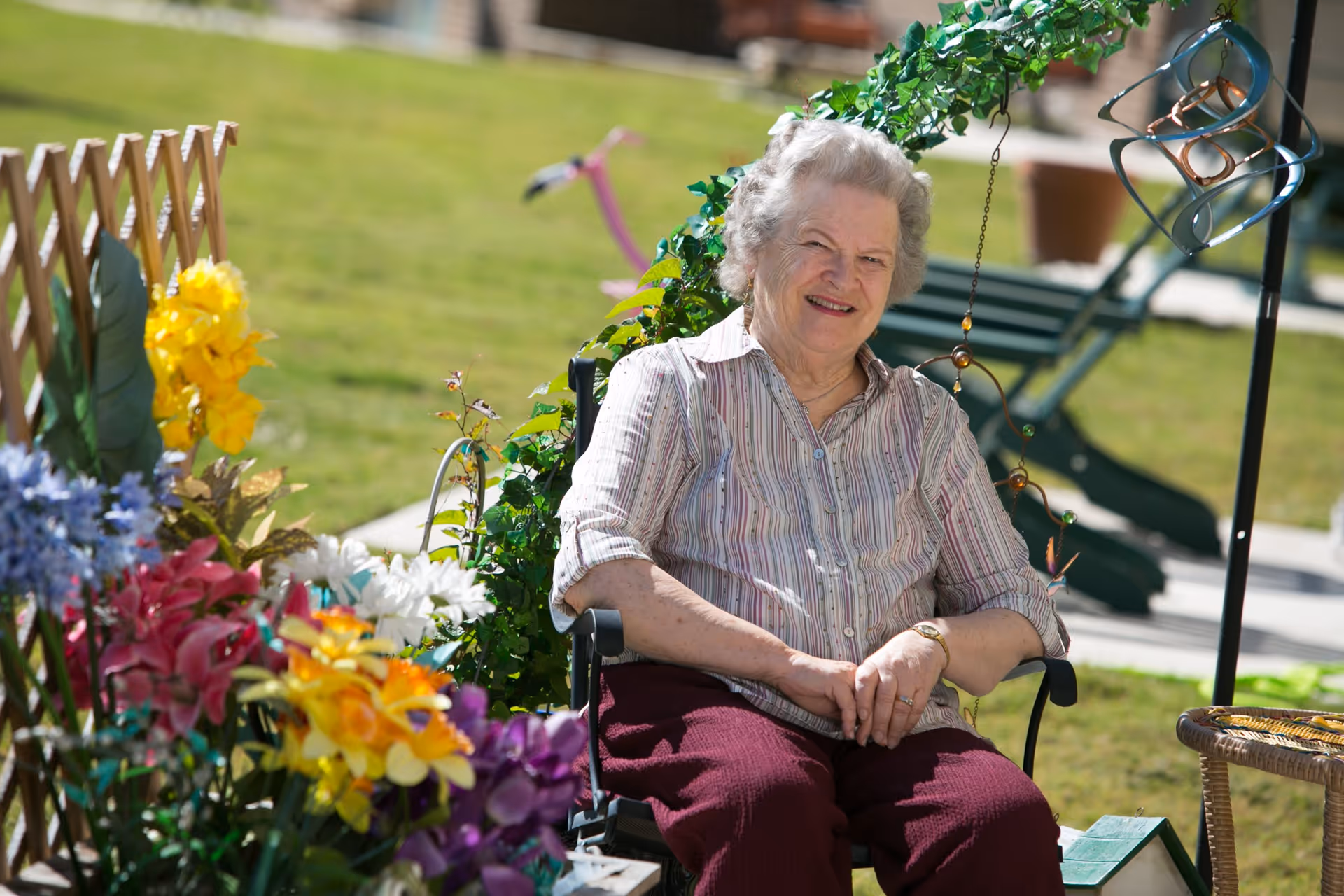 An elderly woman sitting outdoors in a garden area surrounded by colorful flowers and greenery, smiling at the camera. There is a wooden trellis with flowers on the left and a green bench in the background.