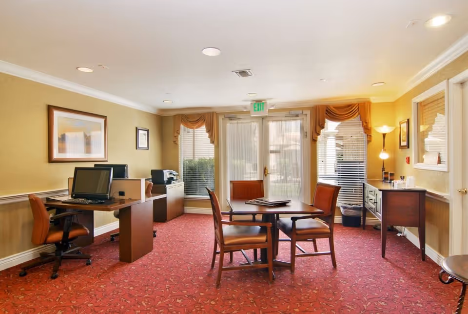 Bright interior common room with desks, computers, a round table and chairs in front of large windows.