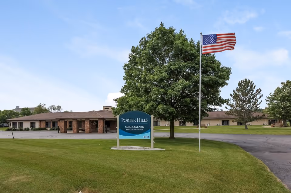 Exterior view of Porter Hills Meadowlark Retirement Village building with a large green lawn, a tree, an American flag on a flagpole, and a sign displaying the facility's name.