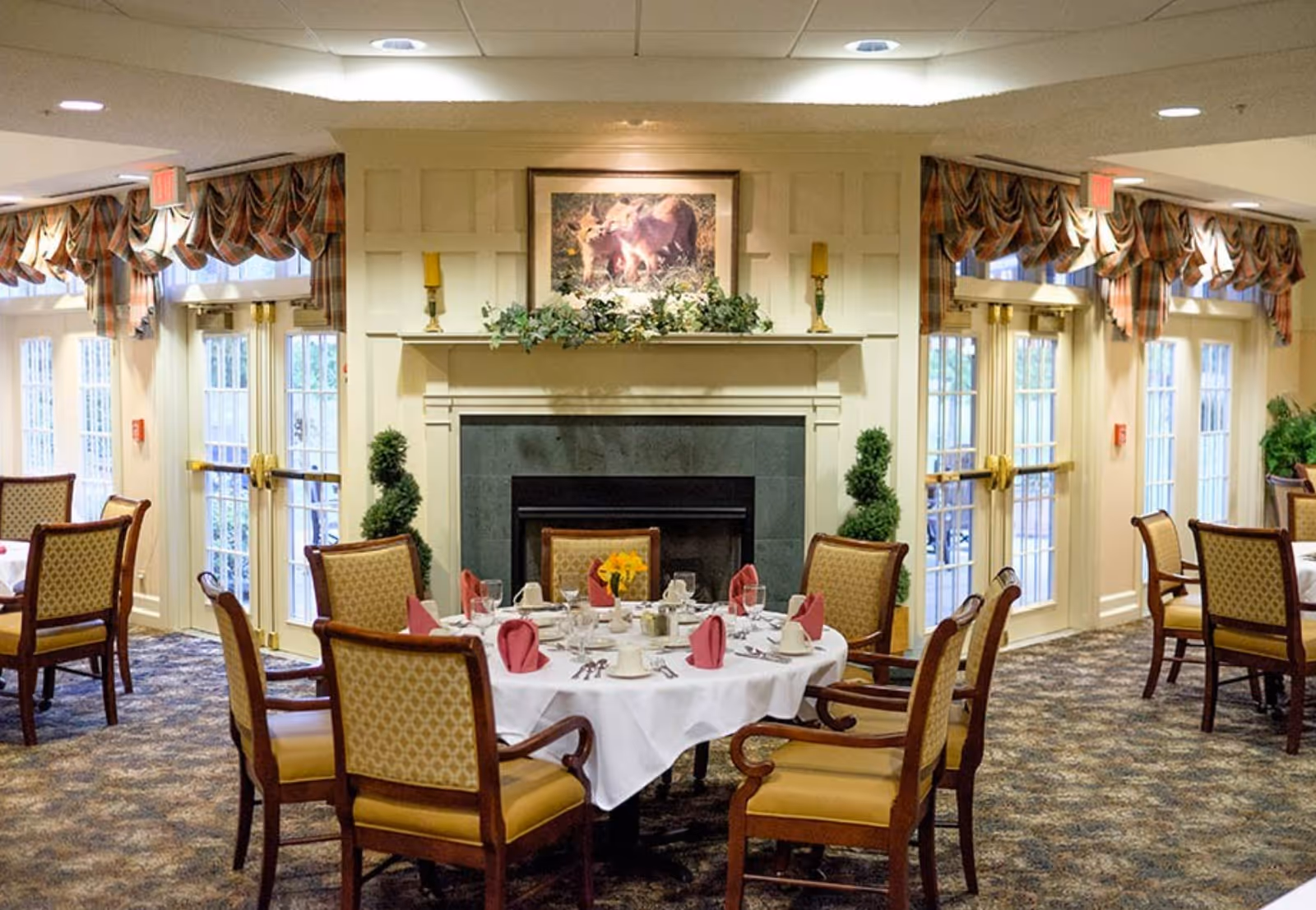 Dining room with round tables set with white linens and folded napkins, upholstered chairs, a fireplace with artwork above, and large windows with draperies.