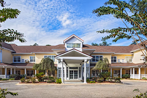 Front exterior view of The Gables of Tallahassee building with a covered entrance, yellow siding, white columns, and landscaped greenery under a partly cloudy blue sky.