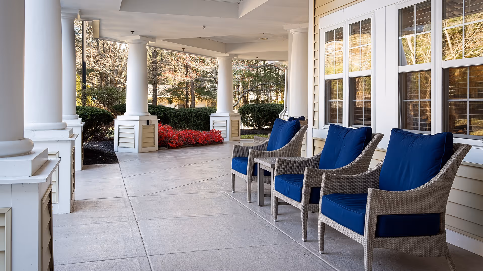 Covered outdoor patio area with three wicker chairs featuring blue cushions and a small table between them, white columns supporting the roof, and bushes and trees in the background.