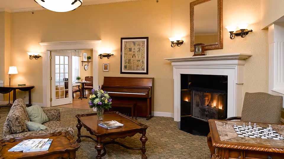A cozy living room with a lit fireplace, a piano against the wall, a coffee table with a vase of flowers, a chessboard on a side table, and comfortable seating including a sofa and an armchair. The room has warm lighting with wall sconces and a large mirror above the fireplace. French doors lead to another room with a table and chairs.