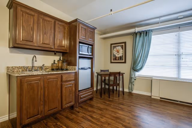A small kitchenette area with wooden cabinets, a granite countertop, a sink, a microwave, and a mini refrigerator. To the right, there is a small wooden table with two chairs, a framed flower picture on the wall, and a window with blue curtains and blinds. The floor is wooden.