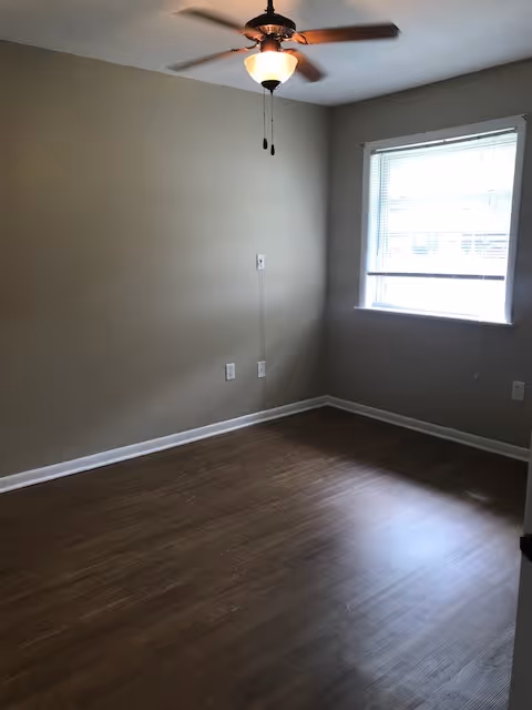 Empty room with wooden floor, beige walls, a ceiling fan with light, and a window with blinds partially open letting in natural light.
