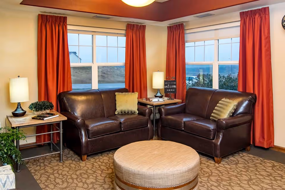 A cozy living room area with two dark brown leather loveseats facing each other, each with a decorative pillow. Between the loveseats is a small wooden side table with a lamp, books, and a small welcome sign. The room has large windows with red curtains and a round beige ottoman in the center on a patterned carpet.
