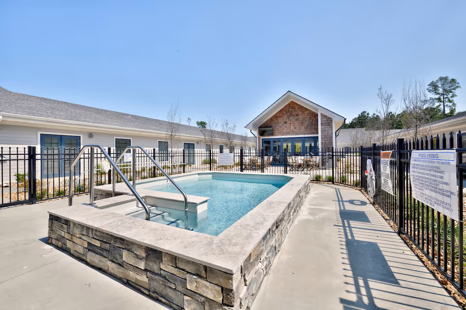 Outdoor swimming pool with stone and concrete edging, surrounded by a black metal fence. Behind the pool is a building with a peaked roof and large windows, with outdoor seating visible. The sky is clear and blue.