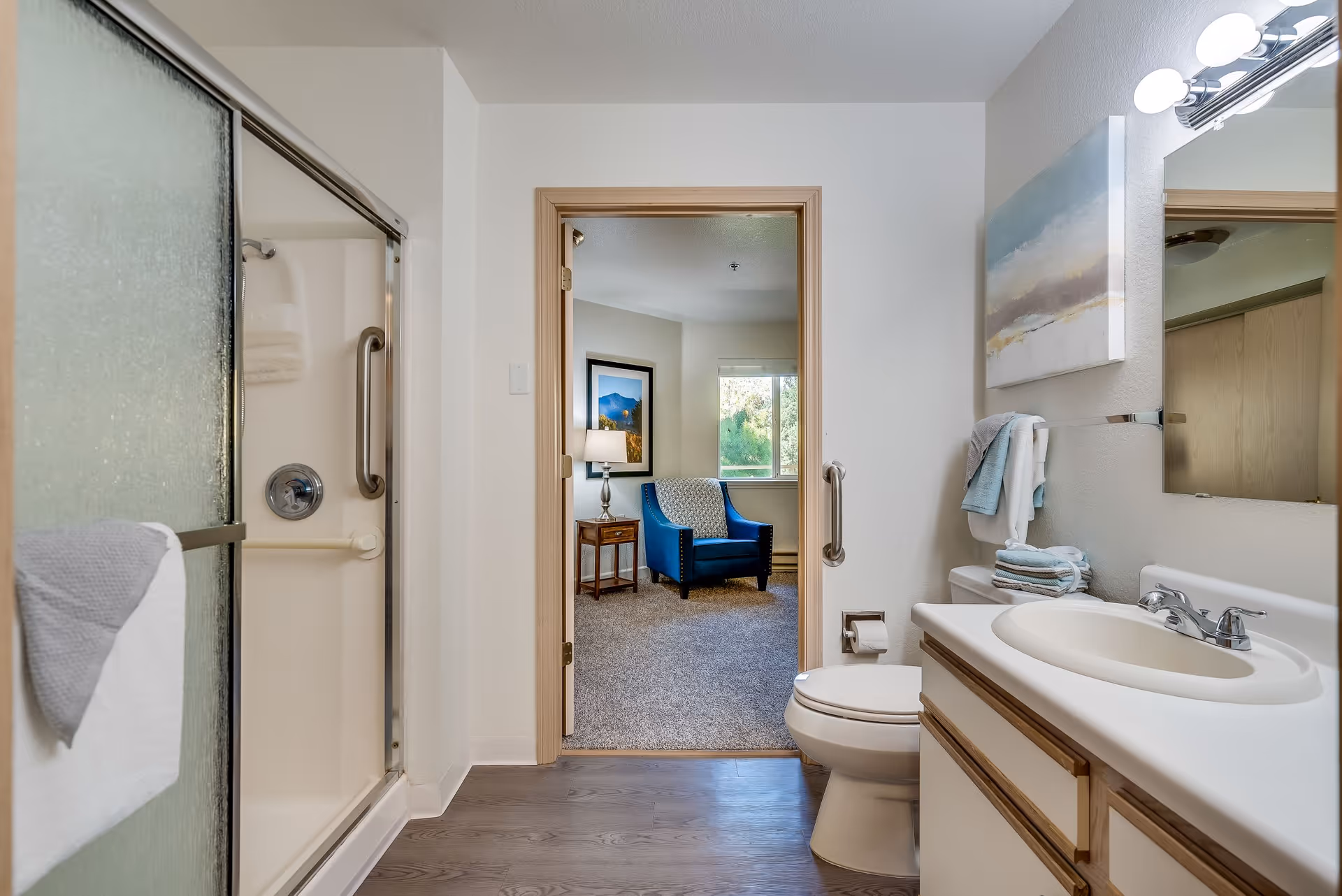 A clean bathroom with a shower featuring frosted sliding doors on the left, a toilet next to a vanity with a sink and mirror on the right. A doorway in the center leads to a carpeted room with a blue armchair, a side table with a lamp, and a window showing greenery outside. Towels are neatly hung and folded on the towel rack and vanity.