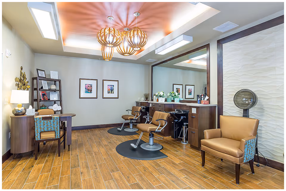 A well-lit salon room with wooden flooring, two salon chairs in front of a large mirror, a hair dryer chair, a small desk with a chair, and decorative lighting fixtures on the ceiling.
