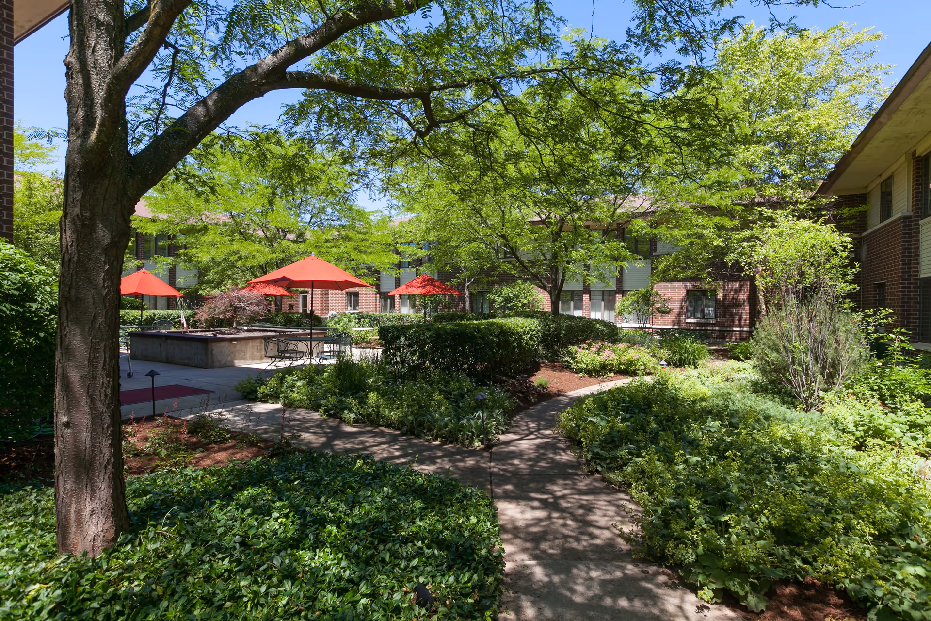 Outdoor courtyard area at Village At Victory Lakes with lush green trees and bushes, a paved walkway, and several tables with red umbrellas. The courtyard is surrounded by brick buildings under a clear blue sky.