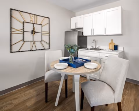 A small dining area with a round wooden table set for two with white plates and a blue tray holding utensils and napkins. Two upholstered beige chairs are positioned around the table. Behind the table is a kitchenette with white cabinets, a stainless steel mini refrigerator, a sink, and a countertop with a cutting board and a blue container. A large decorative wall clock with a metal frame hangs on the light gray wall.