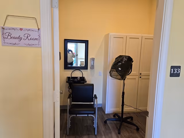 Interior view of a beauty room in a senior living facility. The room features a black salon chair in front of a small black sink with a mirror mounted on the wall above it. To the right, there is a white cabinet and a black hair dryer on a stand. The walls are painted light yellow, and a sign on the door reads 'Beauty Room'. The room number 133 is visible on the door frame.