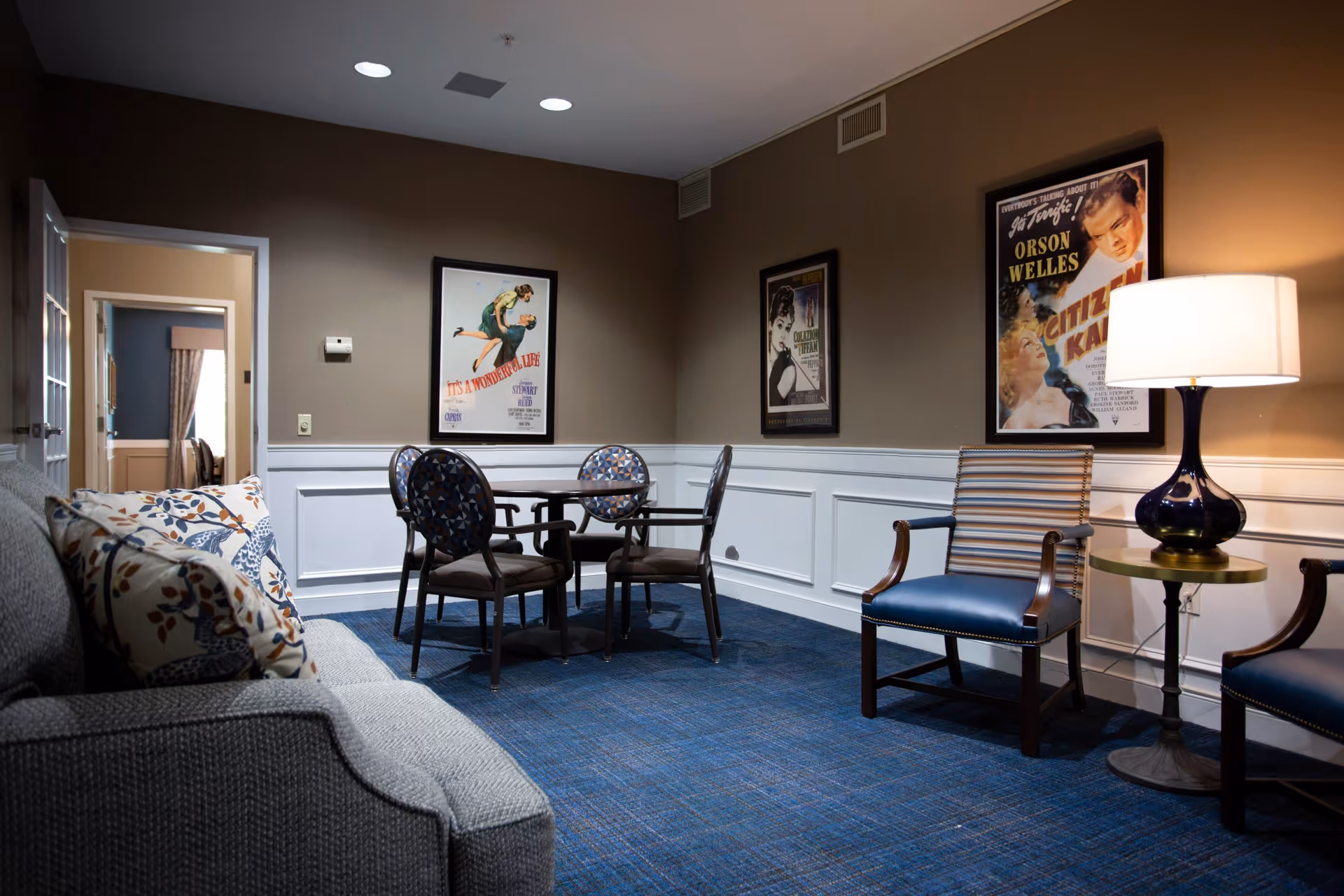 Comfortable common room with a sofa, a small round table and chairs, framed movie posters on the walls, and a floor lamp.