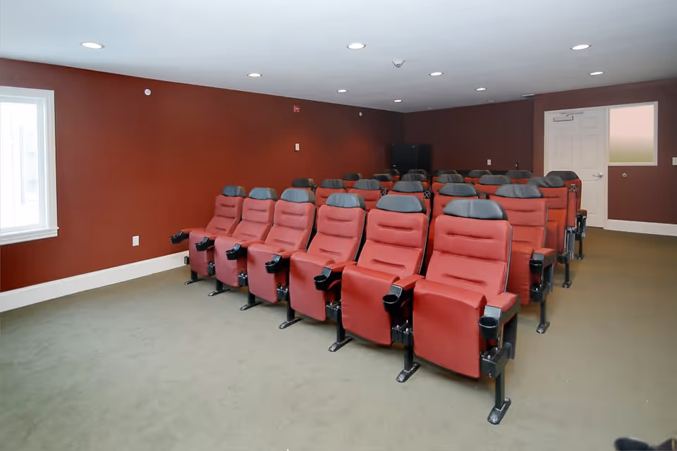 A small theater room with three rows of red cushioned seats with black headrests and armrests, arranged facing a blank wall. The room has maroon walls, a beige carpet, recessed ceiling lights, a window on the left wall, and a white door on the right side.