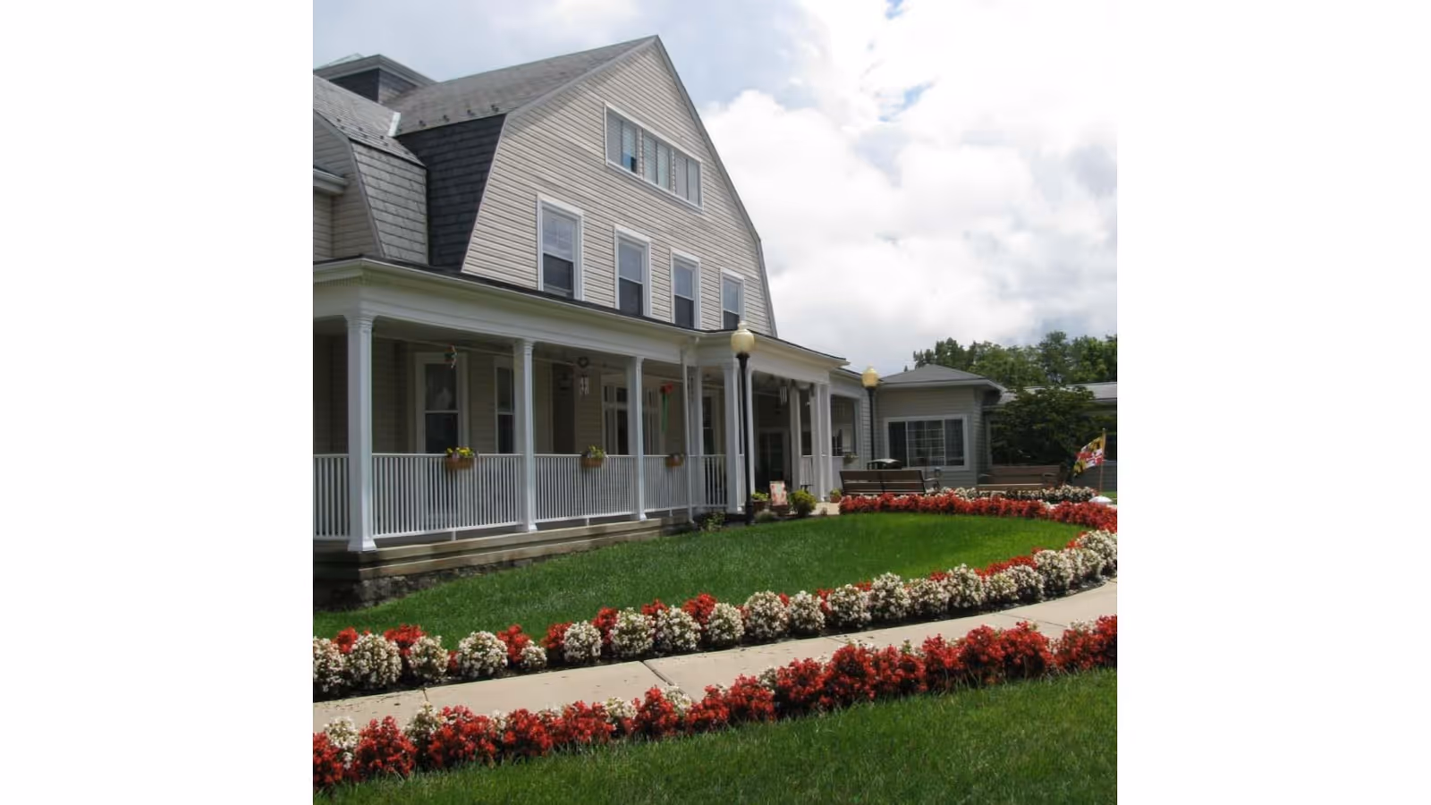 Exterior view of a large, multi-story residential building with a covered porch, white railings, and flower boxes. The foreground features a well-maintained lawn bordered by red and white flowers along a curved sidewalk under a partly cloudy sky.