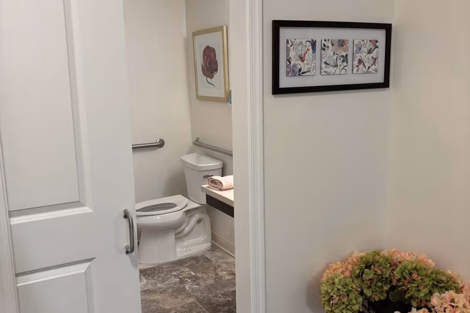 View through a partially open door into a bathroom with a white toilet, grab bars on the walls, a folded towel on the counter, framed floral artwork on the walls, and a flower arrangement in the foreground outside the bathroom.