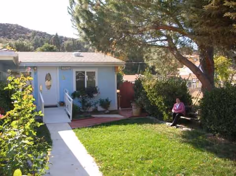 Exterior view of a light-blue senior living home with a resident sitting near greenery.