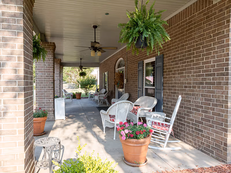 A covered outdoor patio area with brick walls, white wicker chairs with red and white cushions, a white rocking chair, hanging green ferns, potted plants with pink flowers, ceiling fans, and a view of a garden in the background.