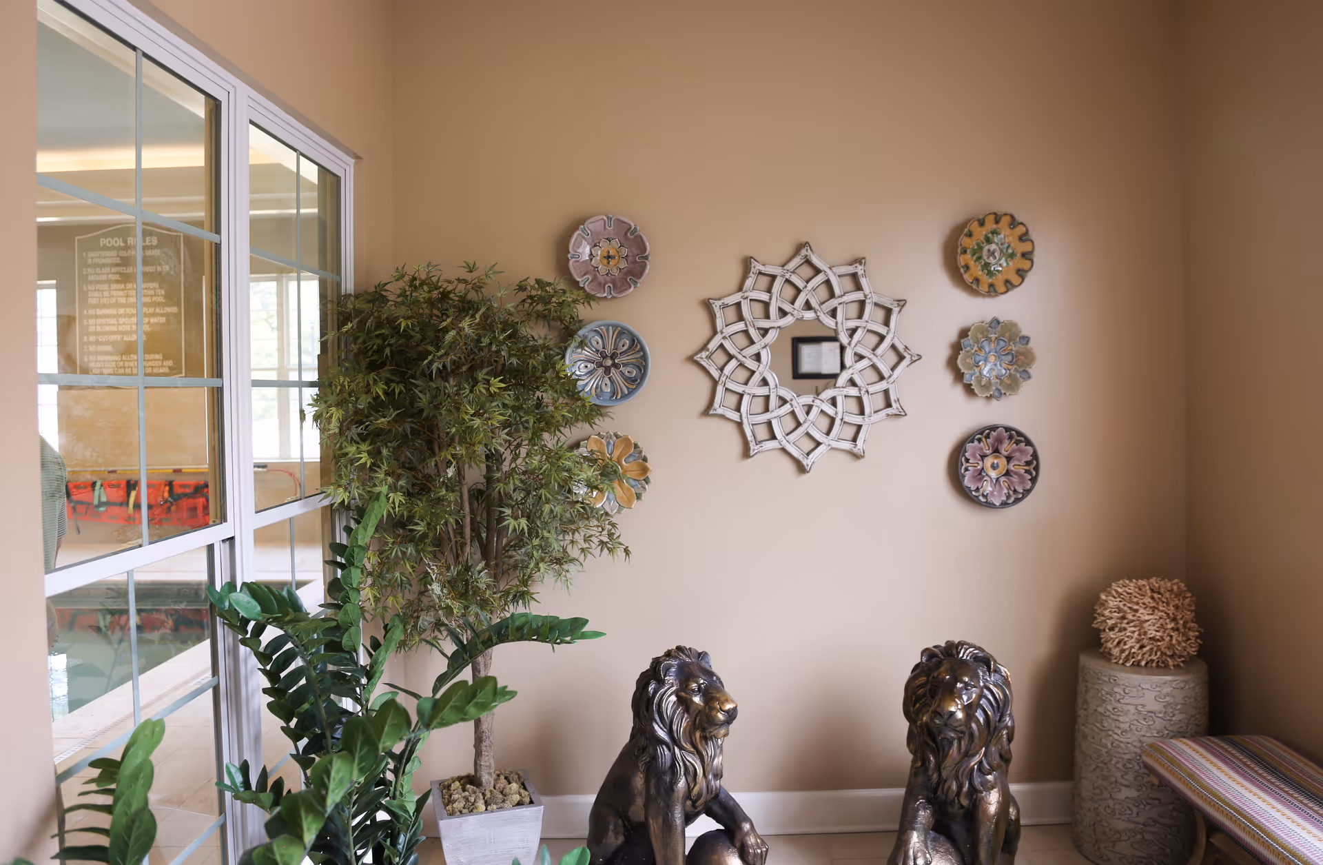 Decorative interior corner with potted plants, wall art, two bronze lion statues and a window looking into an indoor pool area.