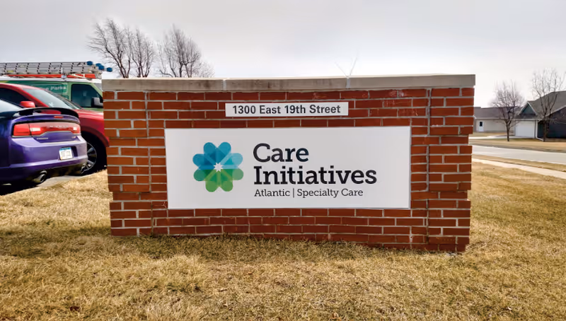 Brick monument sign on a grassy lawn reading "Care Initiatives Atlantic | Specialty Care" with the address "1300 East 19th Street" and parked cars nearby.