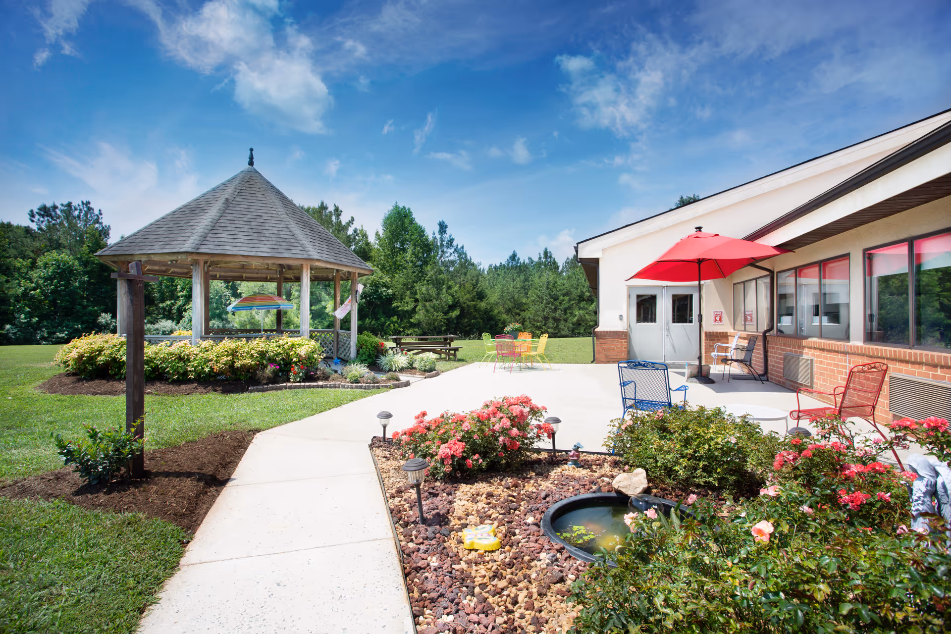 Outdoor patio area at Heritage Hall Brookneal featuring a concrete walkway, a gazebo surrounded by bushes, colorful metal chairs and tables with a red umbrella, and a garden bed with flowers and a small water feature under a bright blue sky.