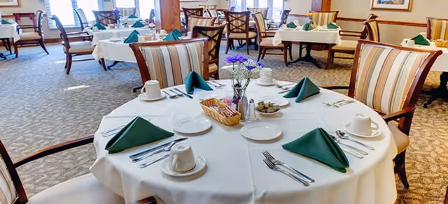 A dining room with round tables covered in white tablecloths, each set with green folded napkins, white plates, cups, and silverware. The chairs have striped upholstery, and there are flowers in small vases on the tables. The room is well-lit with natural light coming through the windows.