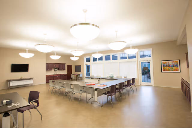 Bright, spacious communal activity/dining room with U-shaped tables and chairs, large ceiling lights, cabinets and floor-to-ceiling windows.