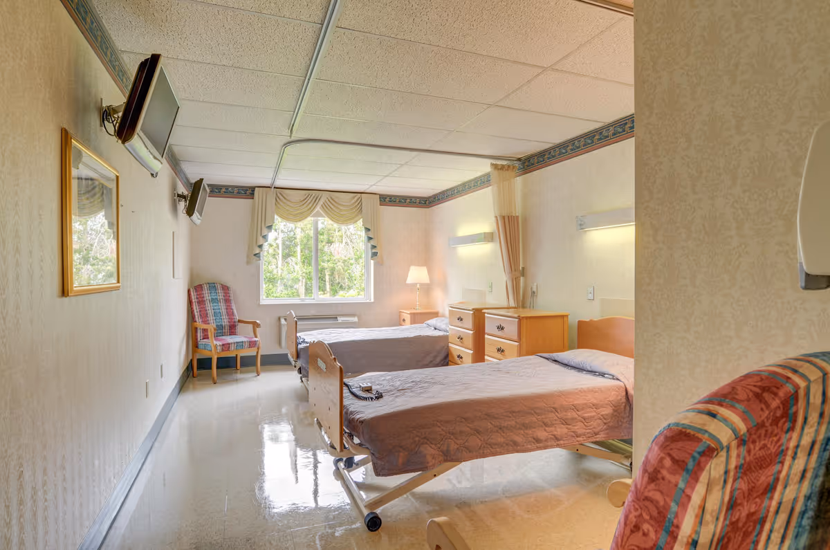 A hospital-style bedroom with two single beds covered in brown quilts, a wooden dresser between the beds, a window with curtains letting in natural light, a plaid armchair near the window, two wall-mounted televisions, and a lamp on the dresser. The room has a clean, clinical appearance with light-colored walls and a tiled floor.